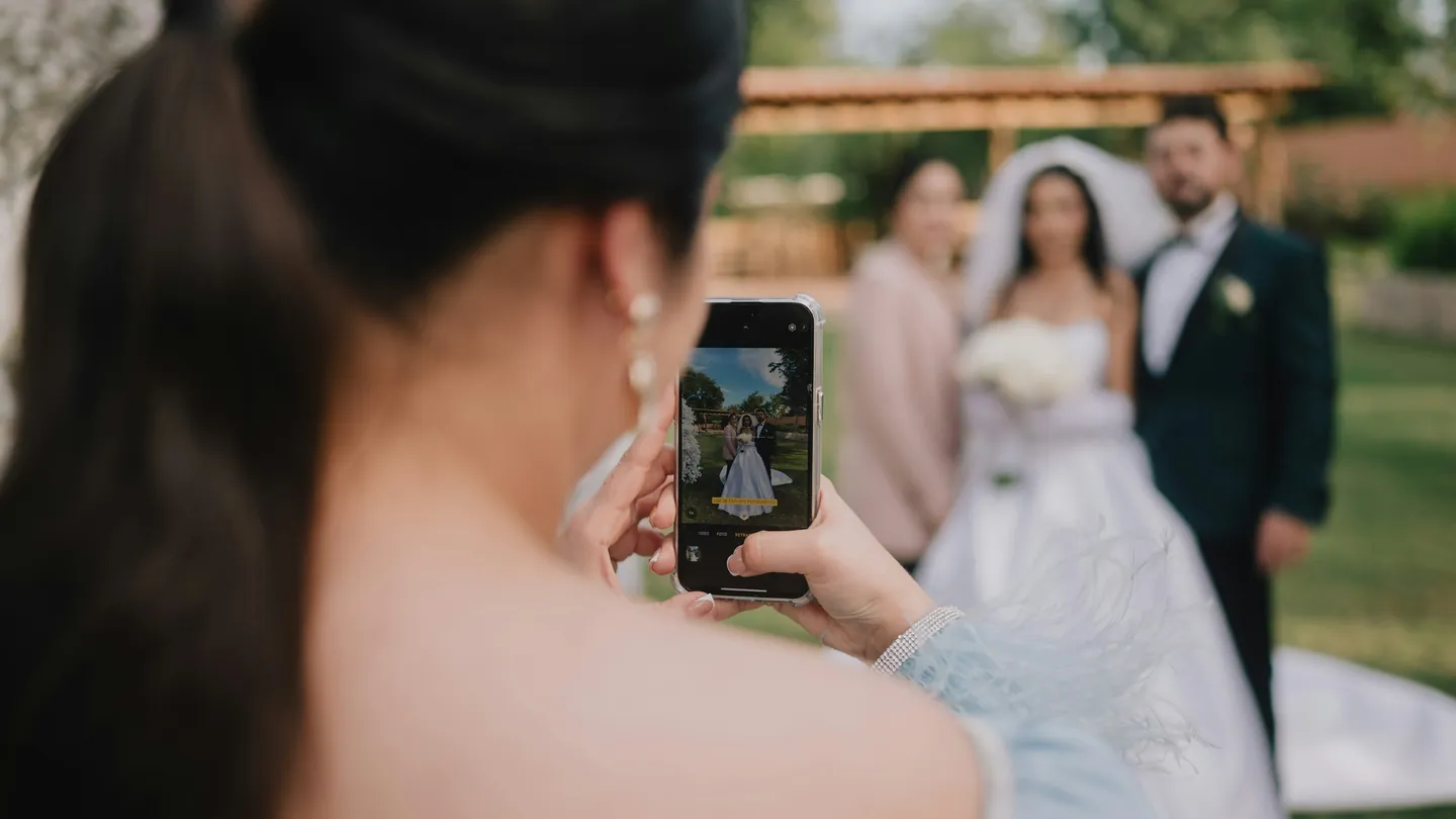 A guest photographs the couple with her phone at an outdoor wedding